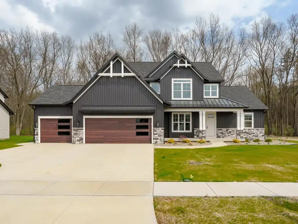 Two-story modern house with dark siding, stone accents, and three-car garage, set on a spacious lot with a concrete driveway and sparse landscaping.