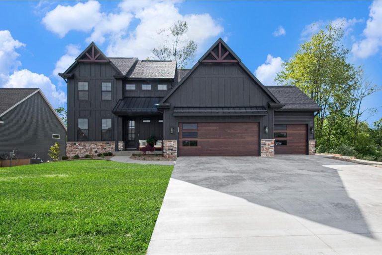 Two-story modern house with dark siding, stone accents, large windows, and a three-car garage, surrounded by a green lawn and trees under a blue sky with clouds.