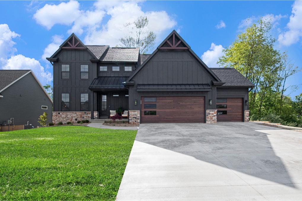 Two-story modern house with dark siding, stone accents, large windows, and a three-car garage, surrounded by a green lawn and trees under a blue sky with clouds.