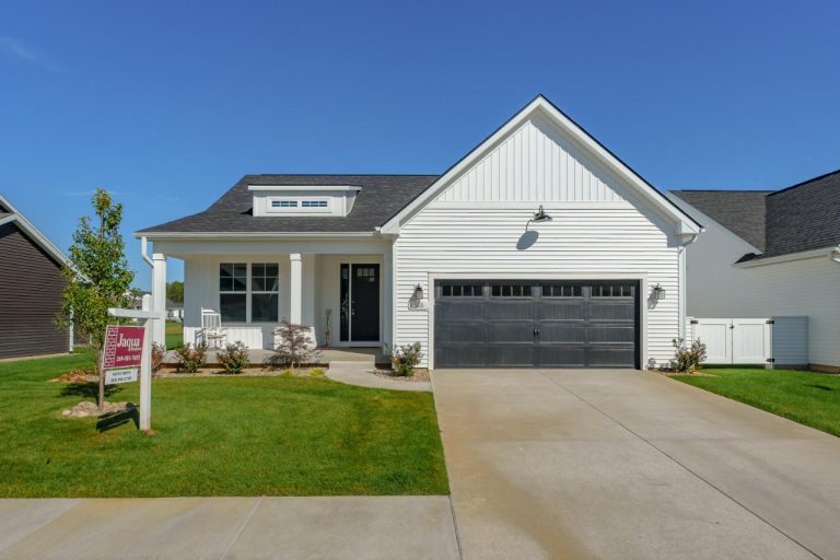 Single-story white house with gray roof, black garage door, and a real estate sign on the lawn. Cement driveway and green grass in front. Clear blue sky in the background.