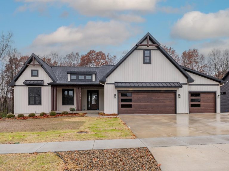 Modern suburban house with a large driveway, double garage, and gray and white exterior, surrounded by trees and a cloudy sky.