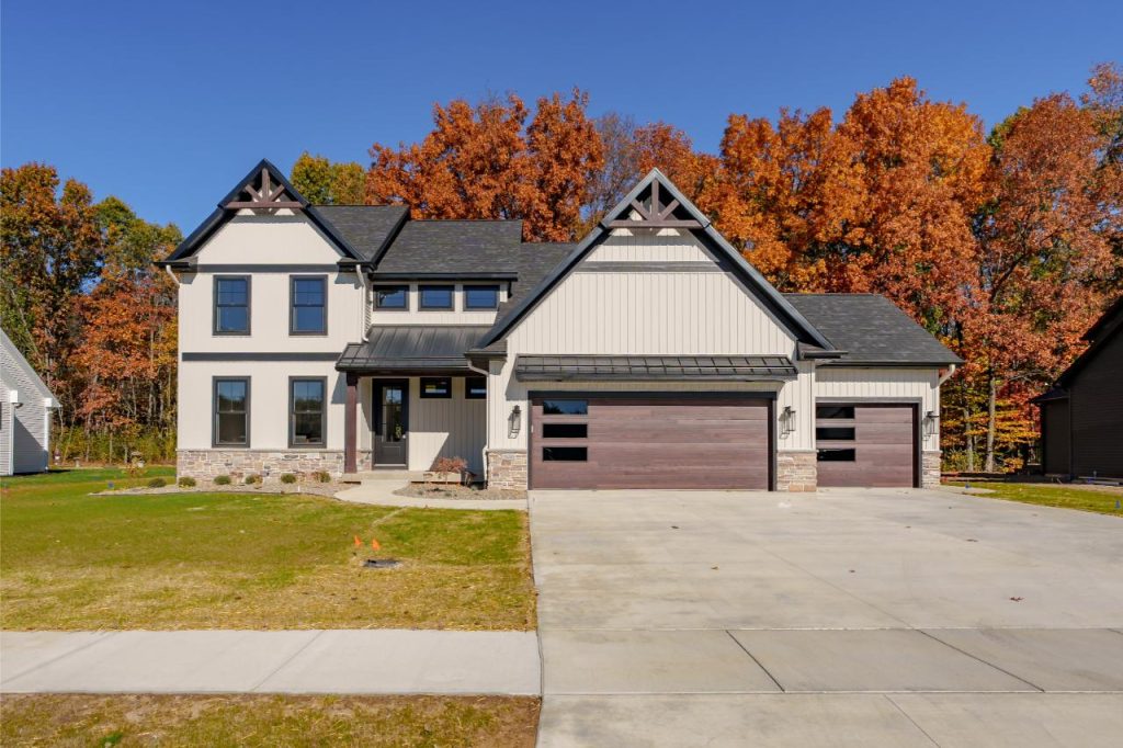 Two-story house with a beige facade, dark trim, and a three-car garage. Surrounded by autumn trees with orange leaves. Concrete driveway and a small lawn in front. Clear blue sky above.