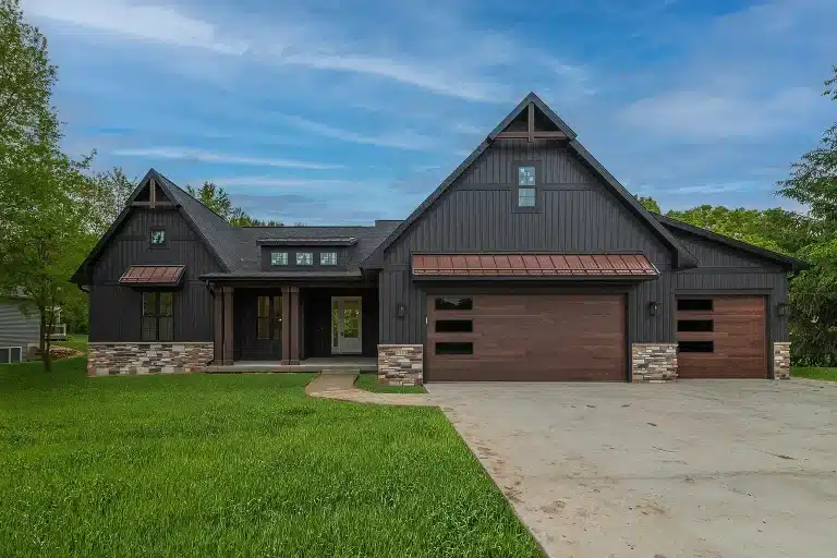 Modern dark gray house with a gabled roof, wooden garage doors, stone accents, and a large driveway, surrounded by green lawn and trees under a blue sky.