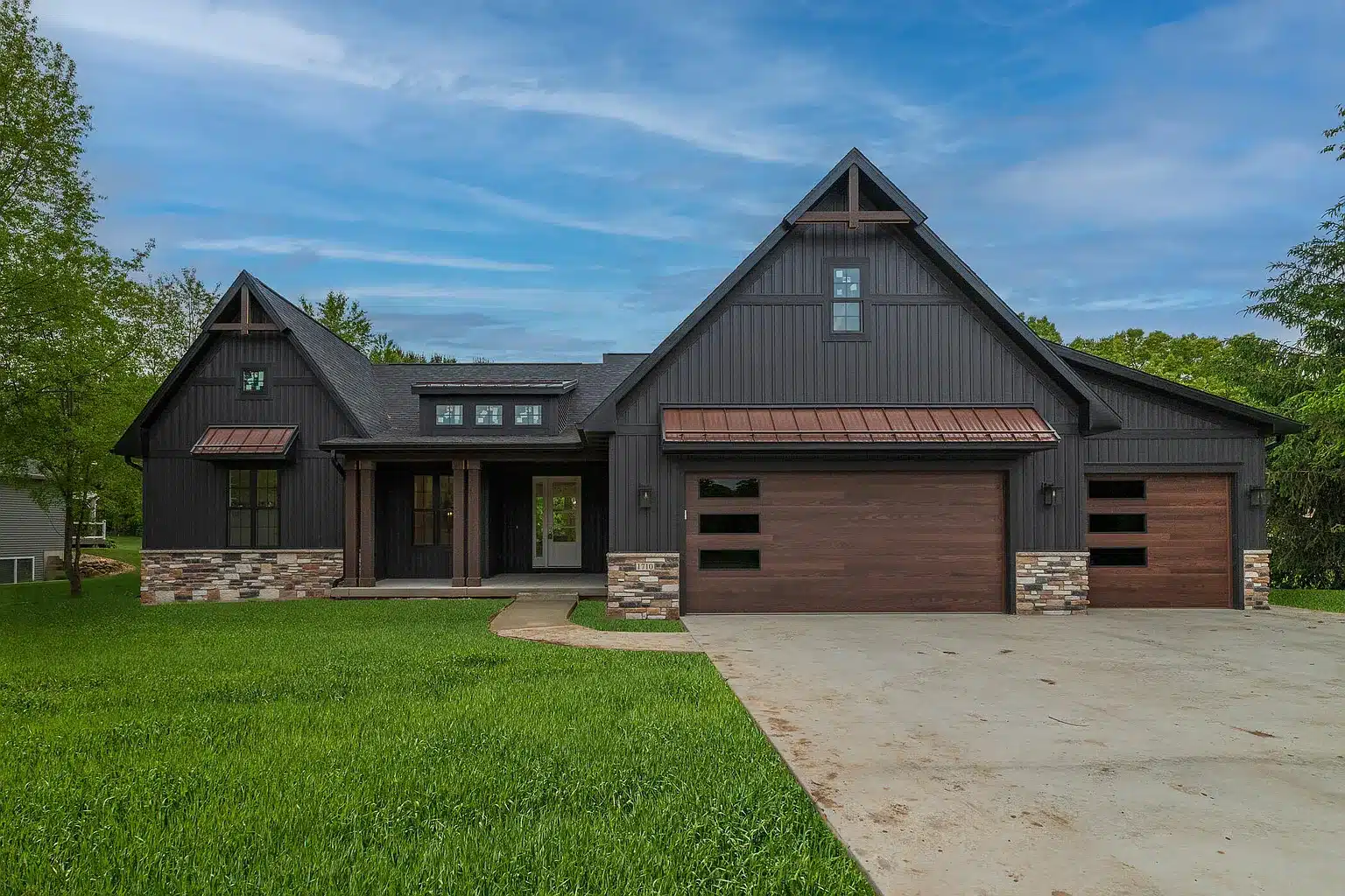 Modern dark gray house with a gabled roof, wooden garage doors, stone accents, and a large driveway, surrounded by green lawn and trees under a blue sky.