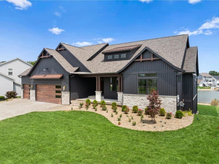 Modern single-story house with dark siding, stone accents, and a three-car garage, set on a green lawn with landscaped shrubs under a blue sky.