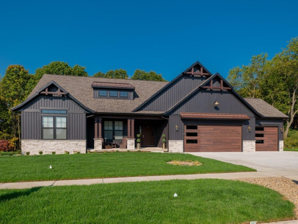 A modern single-story house with dark siding, stone accents, a brown double garage door, and a neatly landscaped front lawn under a clear blue sky.