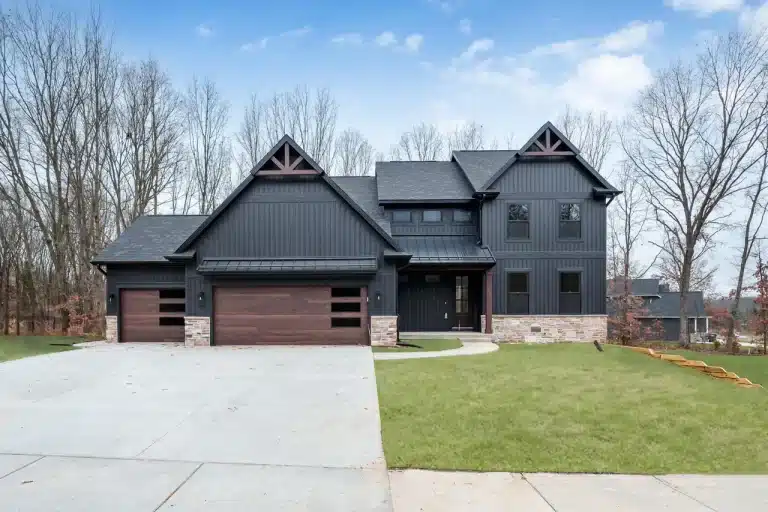 Two-story modern house with dark siding, stone accents, three-car garage with wooden doors, and a large concrete driveway, set against a backdrop of leafless trees and blue sky.