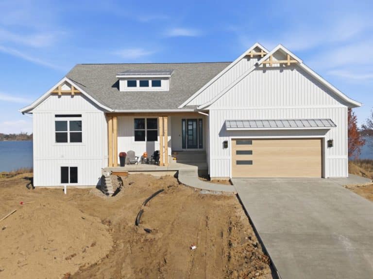 A modern white house with wood accents, a front porch, and an attached garage is under construction by a lake, with unfinished landscaping in the foreground.