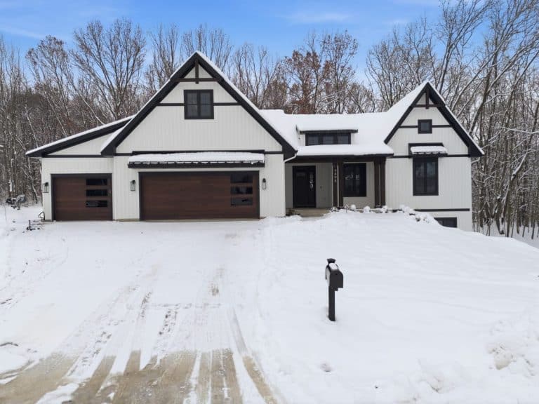 Modern white house with dark trim and a wooden garage door, surrounded by snow, with a snow-covered driveway and mailbox in front. Leafless trees are visible in the background.