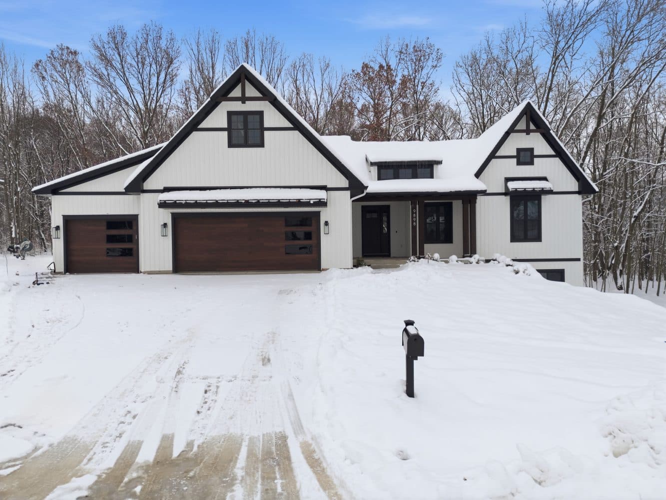 Modern white house with dark trim and a wooden garage door, surrounded by snow, with a snow-covered driveway and mailbox in front. Leafless trees are visible in the background.