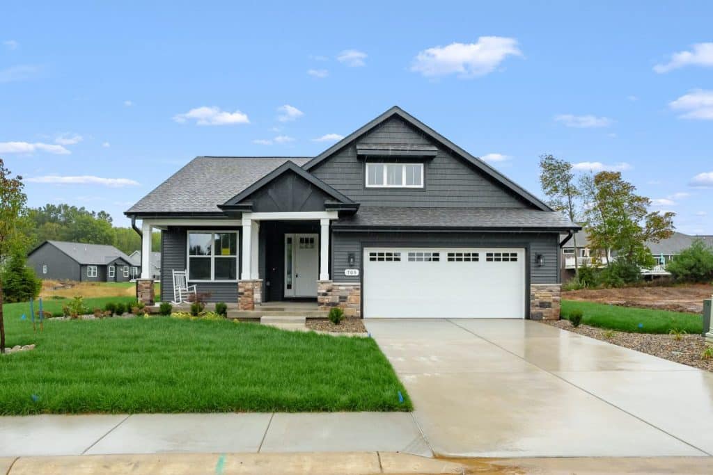 Single-story modern house with dark gray siding, white trim, stone accents, and a two-car garage, set on a green lawn with a concrete driveway under a blue sky.