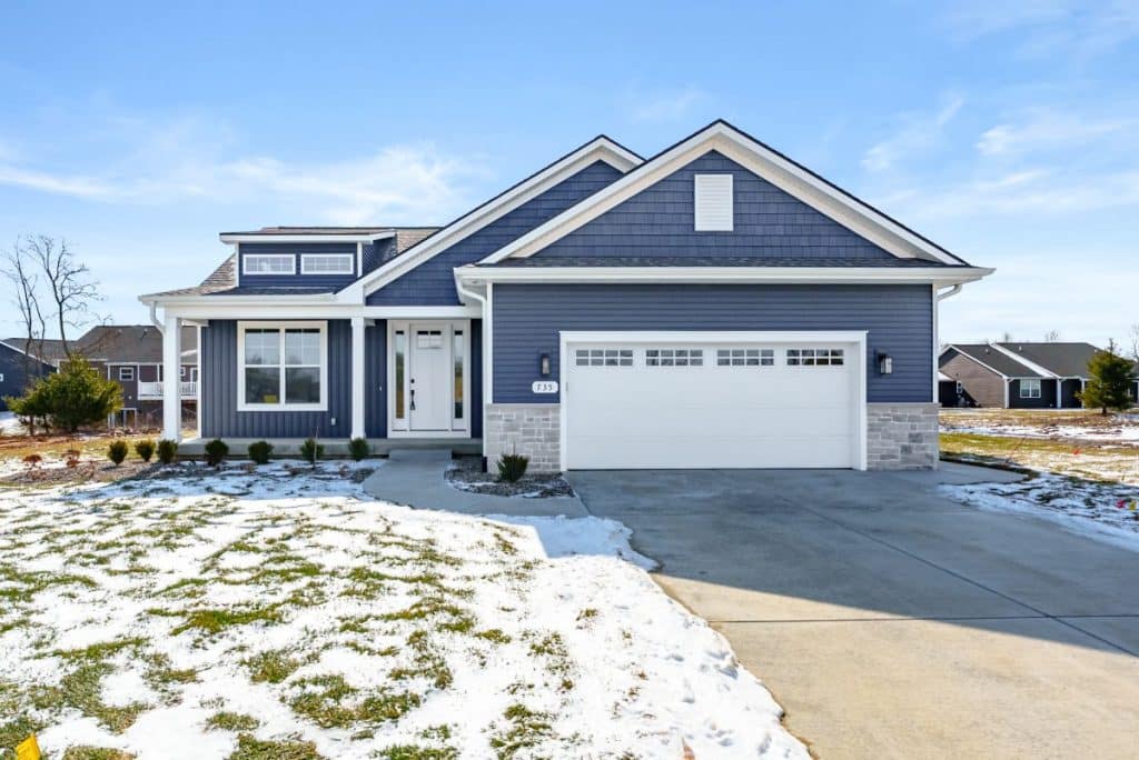 A single-story blue house with white trim, a covered front porch, and a two-car garage, surrounded by patches of snow and grass on a clear day.