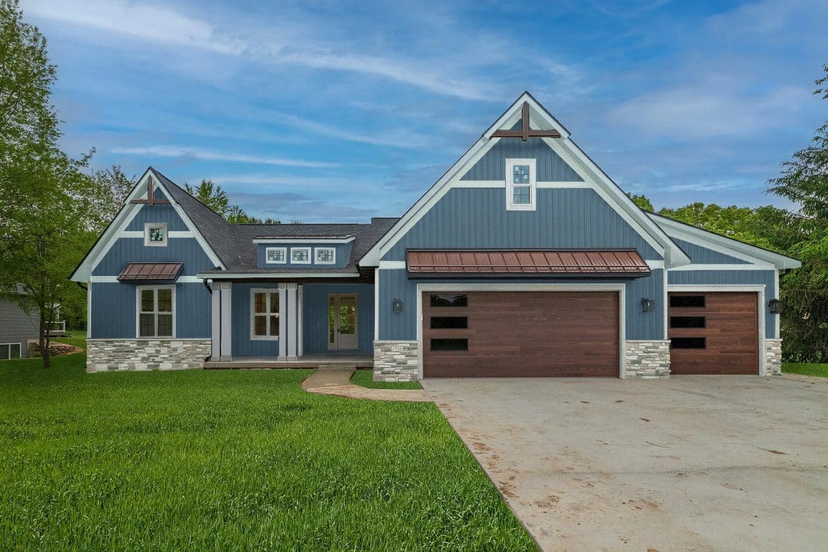 A modern blue house with white trim, stone accents, and two large wooden garage doors, surrounded by green grass and trees under a clear sky.