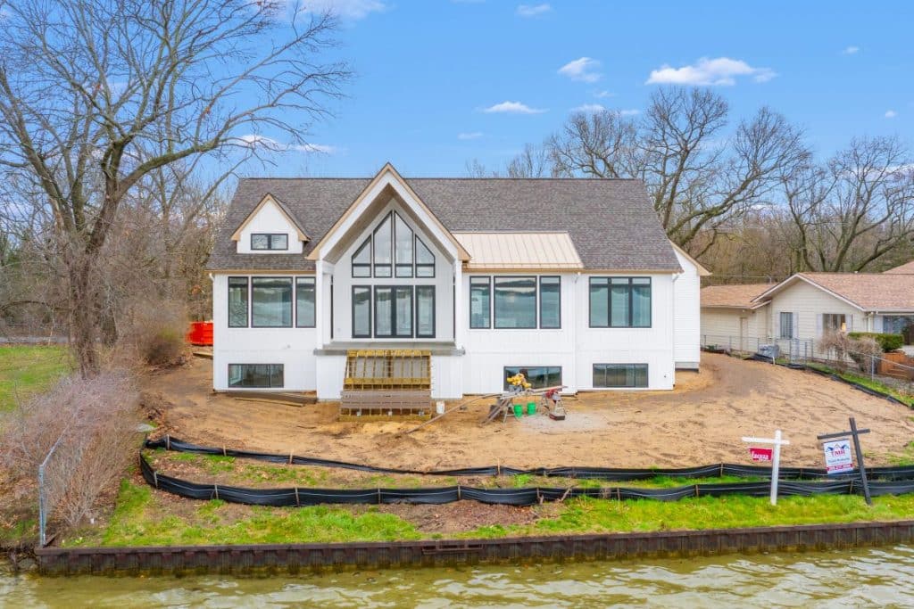 A modern white house with large windows under construction by a lakeshore, surrounded by bare trees and black silt fencing, with construction materials in the yard.