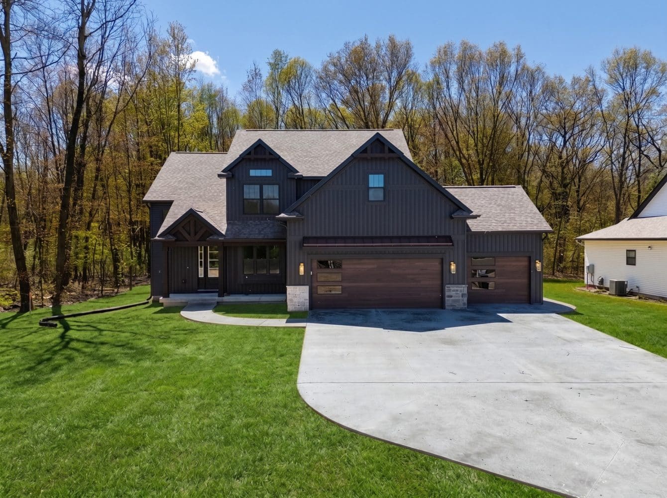 Two-story modern house with dark siding, three-car garage, and stone accents, surrounded by a lawn and trees under a clear blue sky.