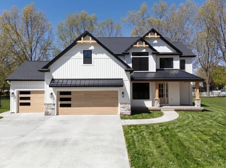 A modern two-story house with white siding, black roof, and wooden accents, featuring a three-car garage and a wide driveway surrounded by green lawn and trees.