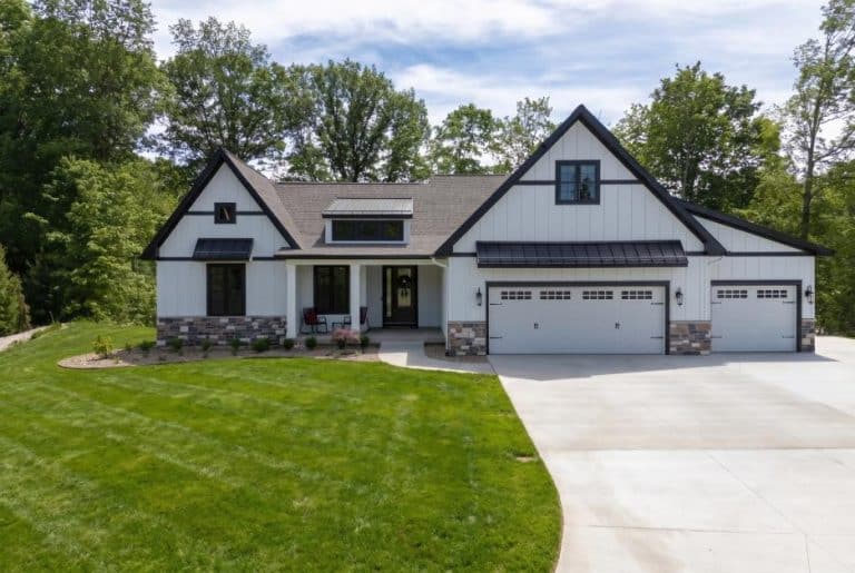 Modern suburban house with white siding, black trim, multiple gables, and a three-car garage, surrounded by green lawn and trees under a partly cloudy sky.