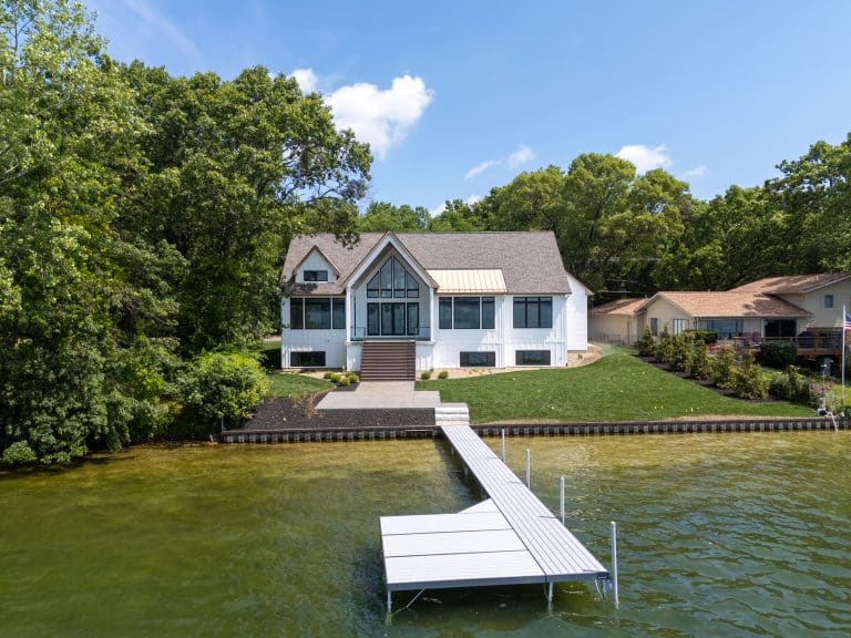 A modern white house with large windows sits by a lake, surrounded by trees, with a private dock extending into the water.