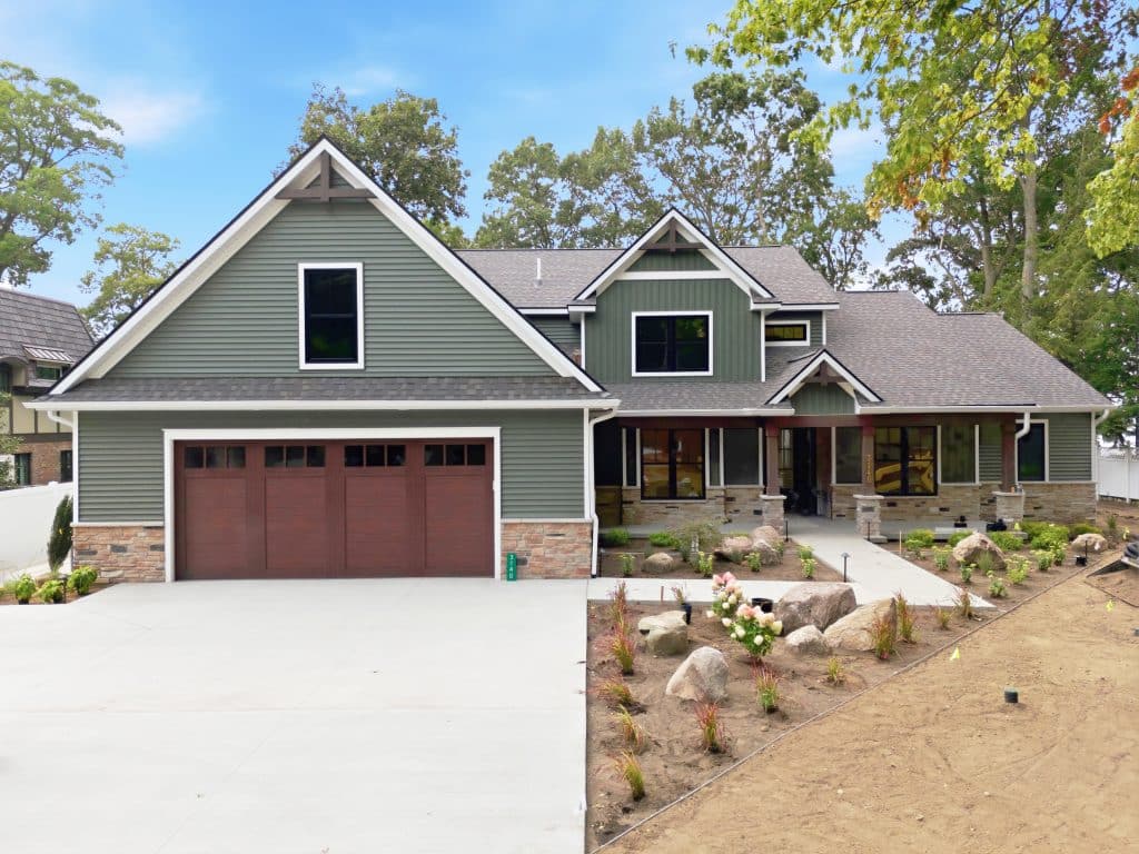 Two-story suburban house with green siding, stone accents, a covered front porch, and double wooden garage doors, surrounded by a landscaped yard and trees.