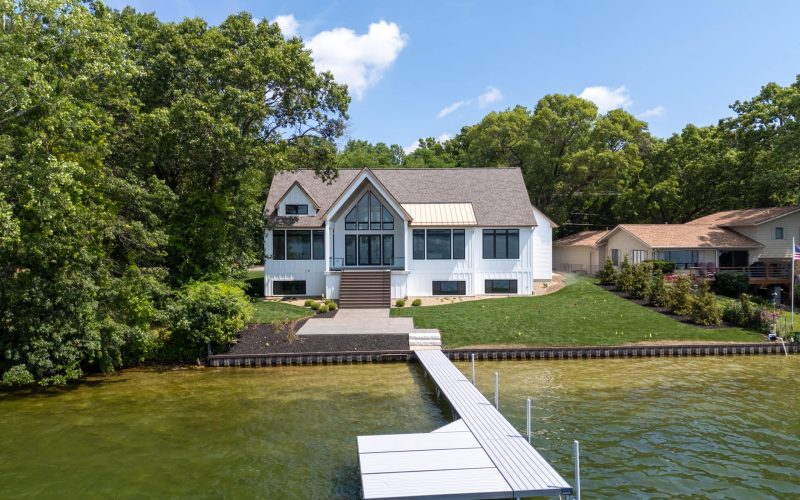 A modern white house with large windows sits by a lake, surrounded by trees, with a private dock extending into the water.