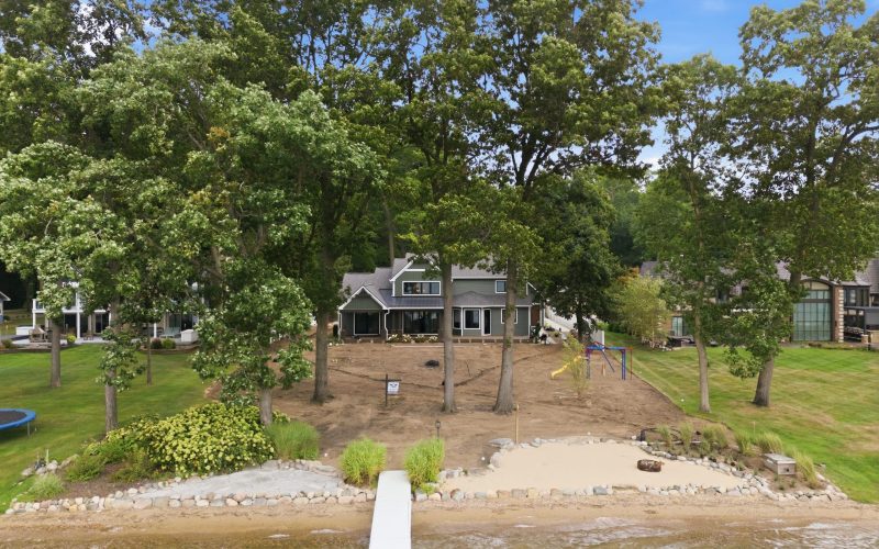 Two-story lakefront house with large trees in the backyard, sandy area near the water, dock extending into the lake, and playground set to the right.