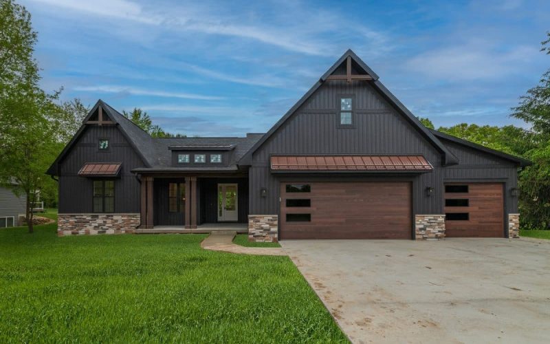 A modern dark gray house with a large driveway, wooden garage doors, stone accents, and a green lawn under a blue sky.