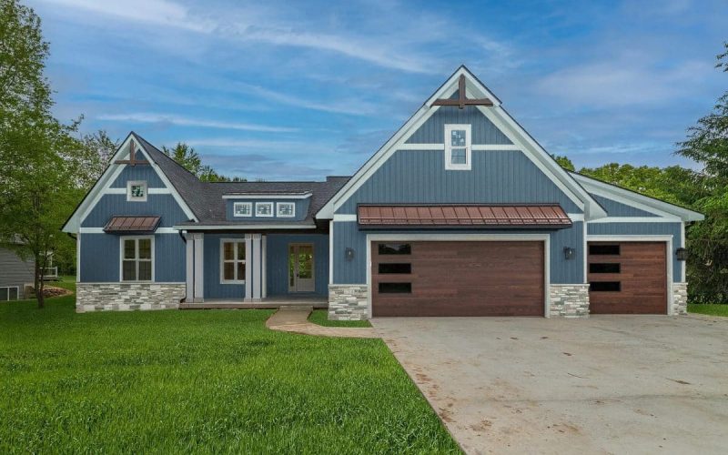 A modern blue house with white trim, stone accents, and two large wooden garage doors, surrounded by green grass and trees under a clear sky.