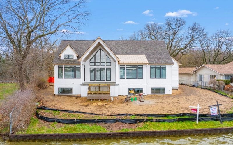 A modern white house with large windows under construction by a lakeshore, surrounded by bare trees and black silt fencing, with construction materials in the yard.